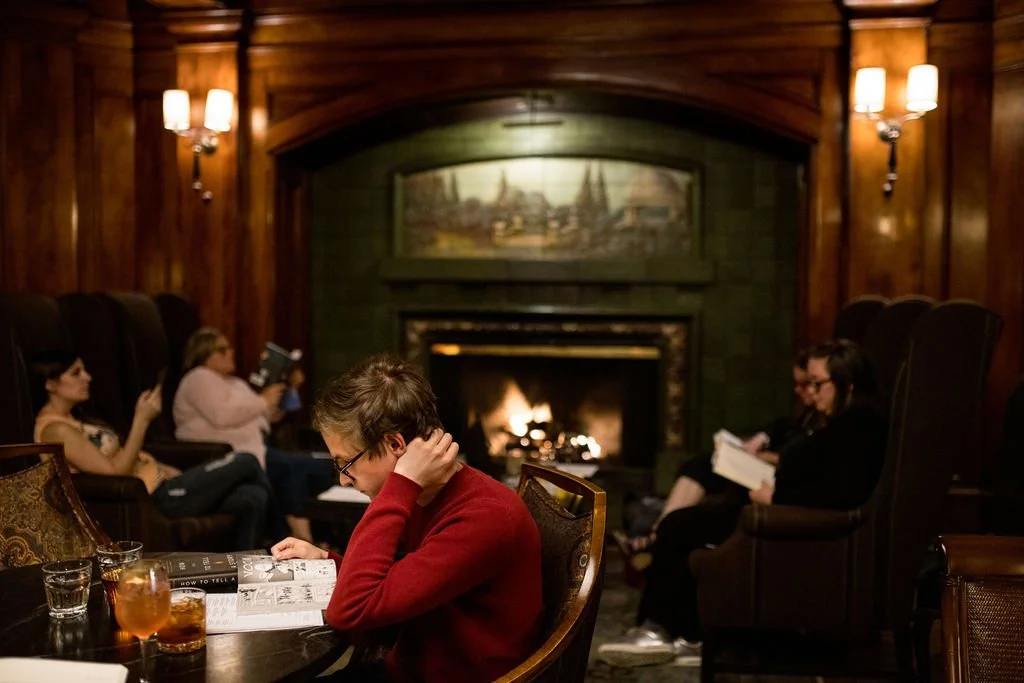 Image of a wood-paneled room with a lit fireplace and five people quietly reading.